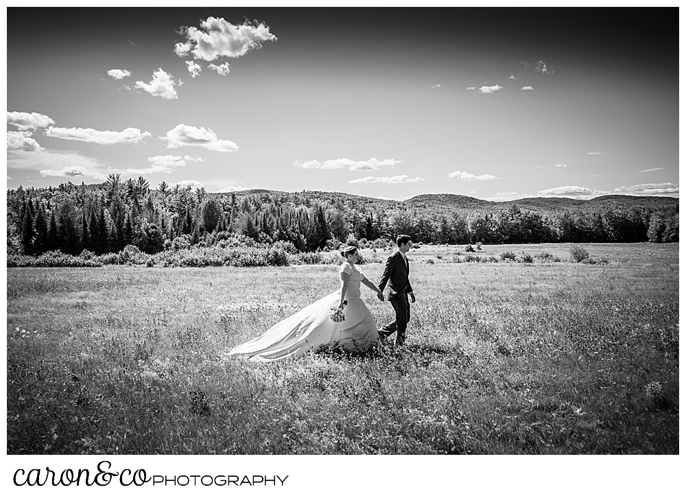 black and white photo of a bride and groom holding hands, walking in a field, with mountains in the background, during a sweet western Maine wedding