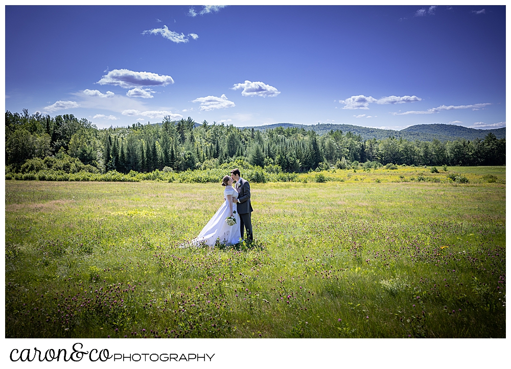 a bride and groom stand in a field, kissing, the mountains in the background, of a sweet western Maine wedding