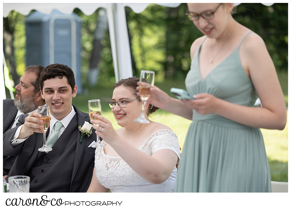 a bride and groom raise their champagne glasses for a toast while a bridesmaid reads her phone