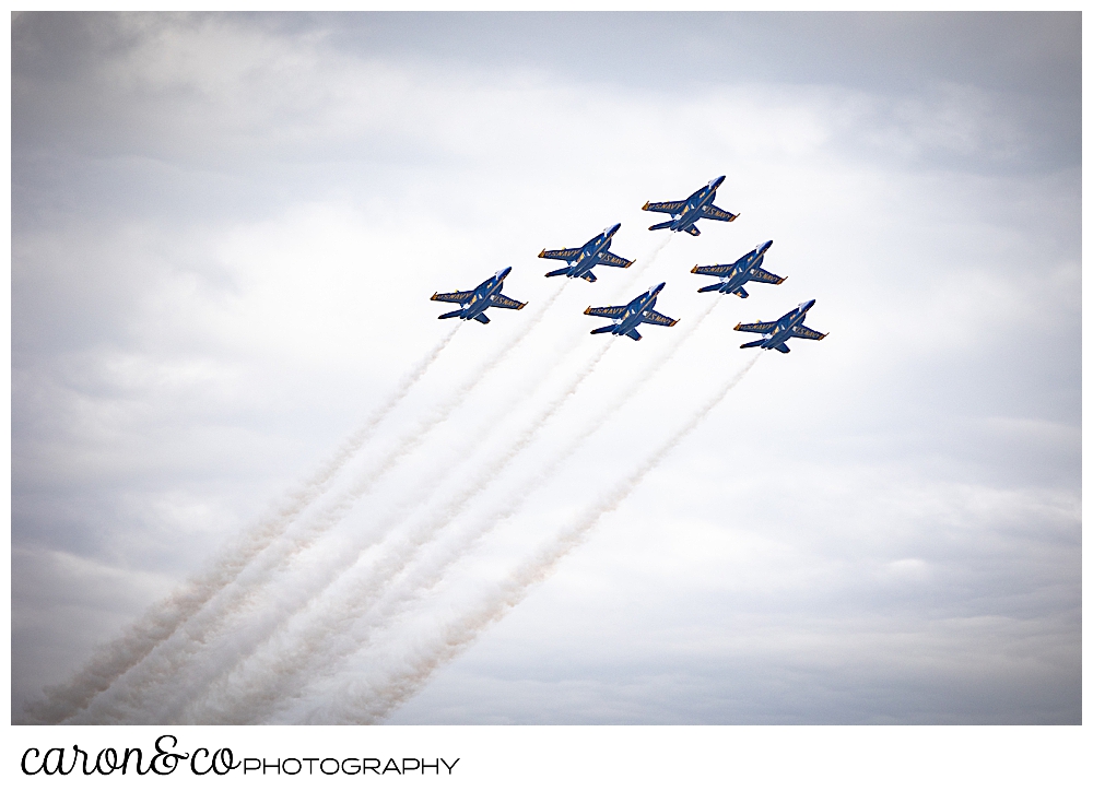 Flying in a diamond are 6 us navy blue angels, with contrails following through the clouds