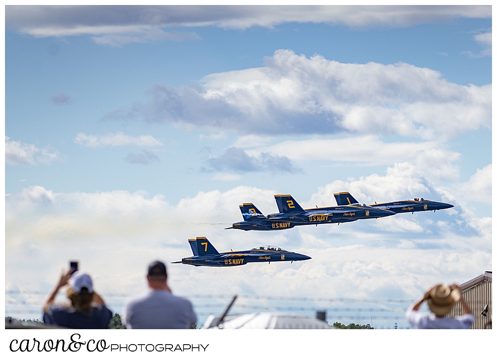 4 us blue angels airplanes flying low in the background, as spectators watch and take photos in the foreground