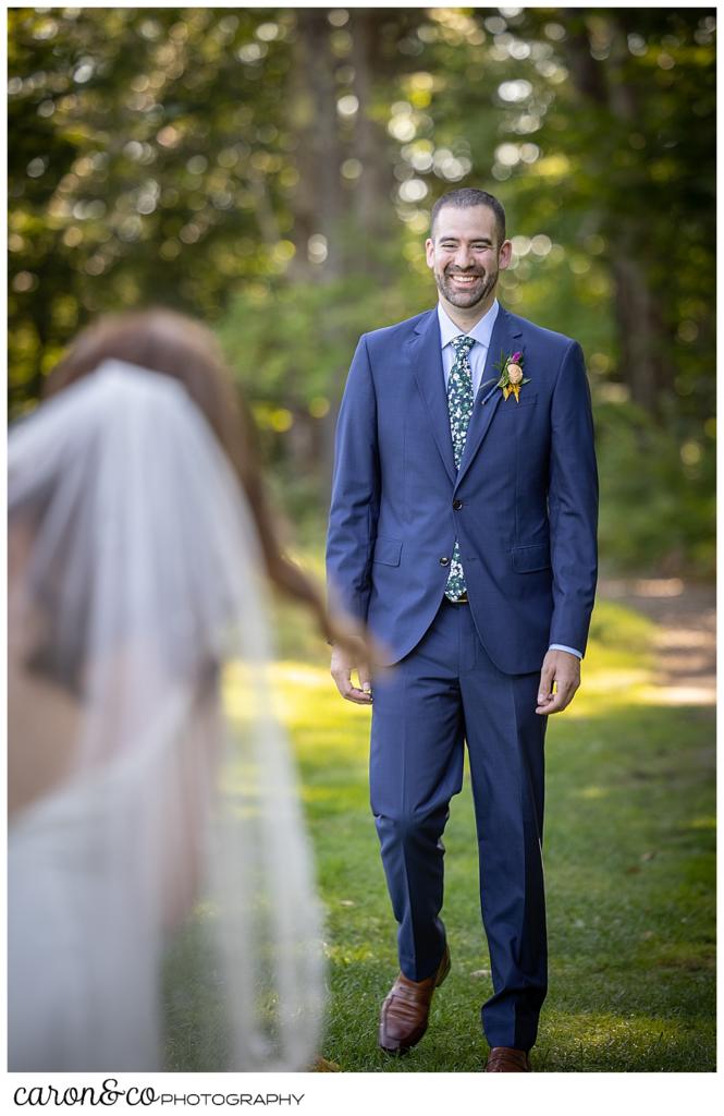 a groom wearing a blue suite, smiles and walks toward his bride in the foreground