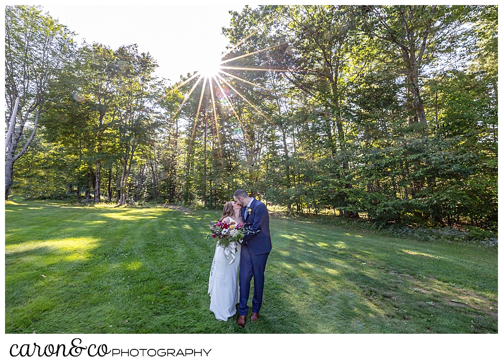 a bride and groom kissing in a grassy field, surrounded by trees