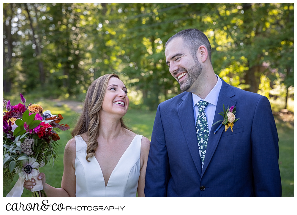 a bride and groom look at one another and smile, the bride is holding her bouquet of fall flowers