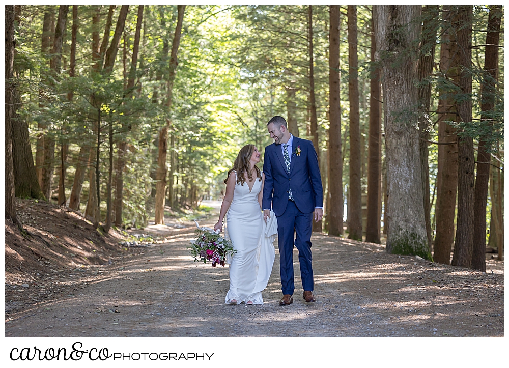 a bride and groom walk hand in hand down a dirt road lined with tall trees