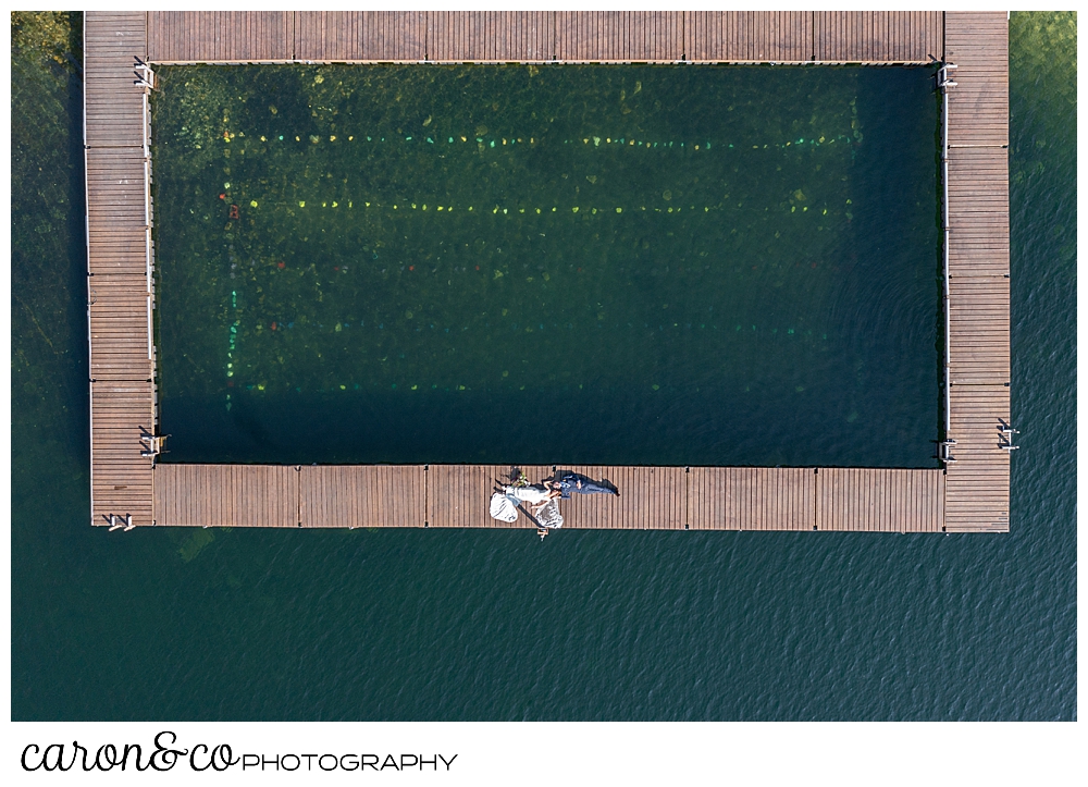 a drone photo of a bride in a white dress, and a groom in a blue suit, lie on a dock at Camp Skylemar on Trickey Pond in Naples, Maine