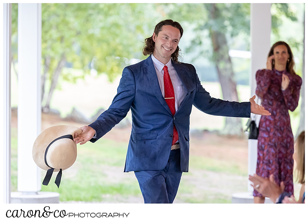 a wedding guest in a blue suit and red tie, holds a straw hat in one hand