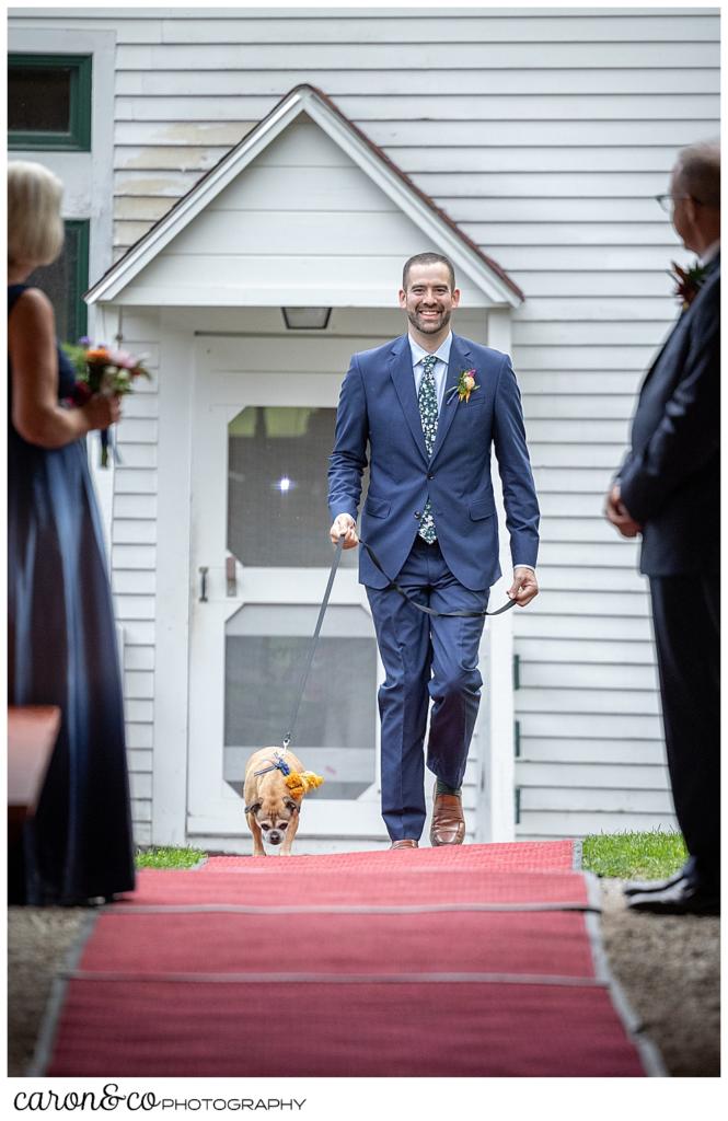 a groom wearing a blue suit, with a small dog on a leash, walks down a red carpet toward the ceremony site under the pavilion at Camp Skylemar