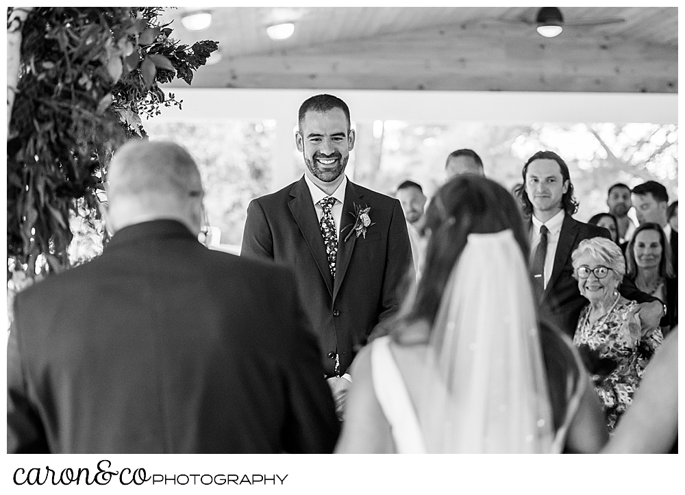 a black and white photo of a smiling groom in the background, and the backs of the bride and her father