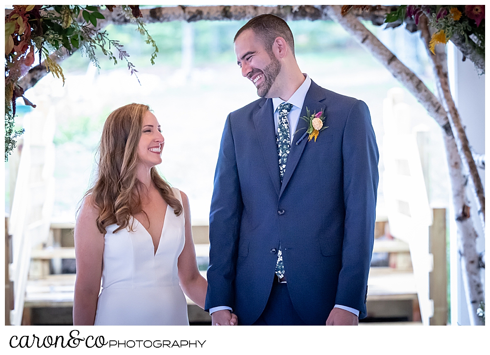 a smiling bride and groom stand under an arbor at their Camp Skylemar wedding ceremony