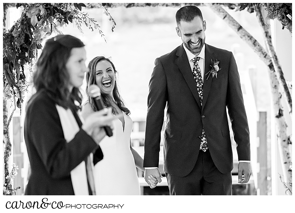 a black and white photo of an officiant holding a microphone in the foreground, and a laughing bride and groom holding hands in the background