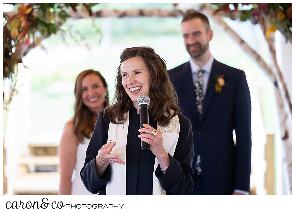 a reverend who is smiling and holding a microphone is in the foreground, a smiling bride and groom are behind her