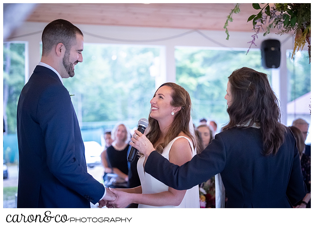 a bride and groom are standing facing one another, holding hands, the bride is smiling at the groom, as the Reverend holds a microphone for the bride to speak her vows