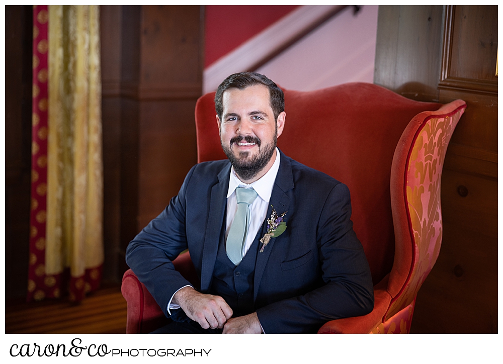 a groom portrait, of a dark haired, bearded groom sitting in a red chair