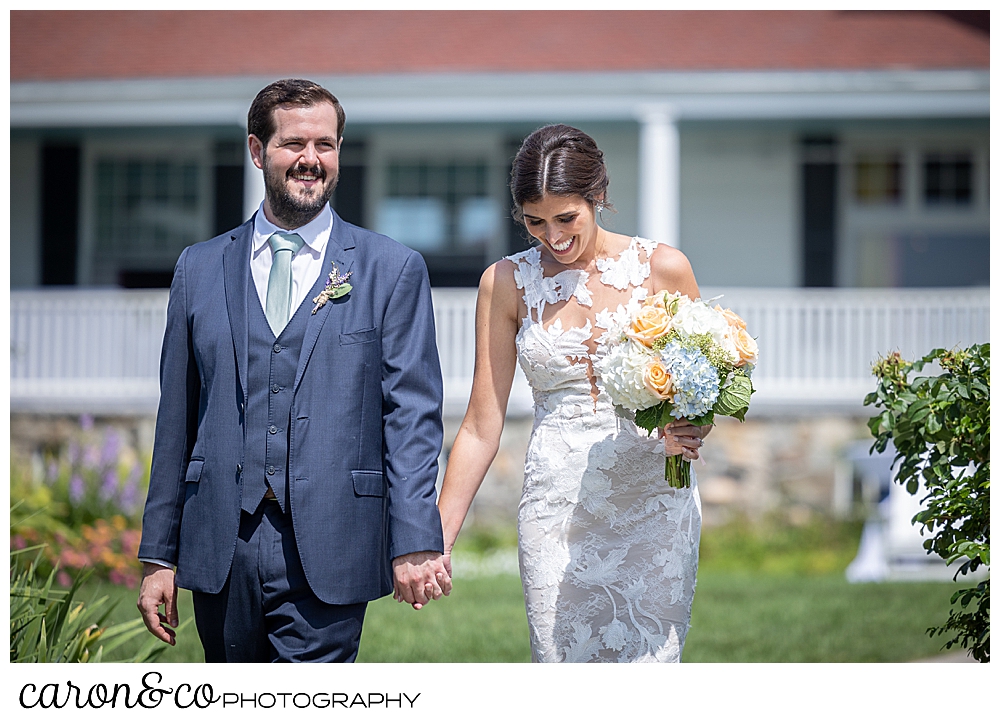 a bride and groom walking on the lawn of the Colony Hotel, the bride is looking down while she holds a bouquet of white, pink, blue, and green