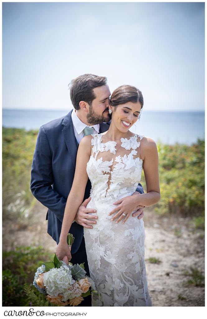 a dark-haired bride and groom stand in a loose embrace, the groom behind the bride, nuzzling her ear