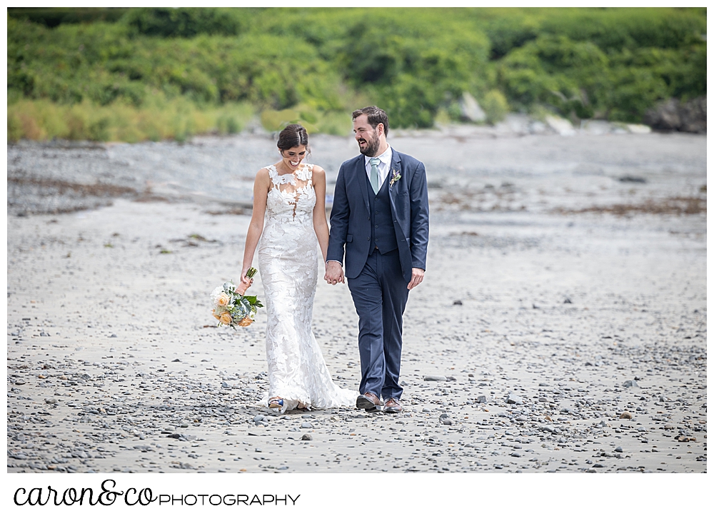 a dark-haired bride and groom hold hands and walk on Colony Beach in Kennebunkport, Maine