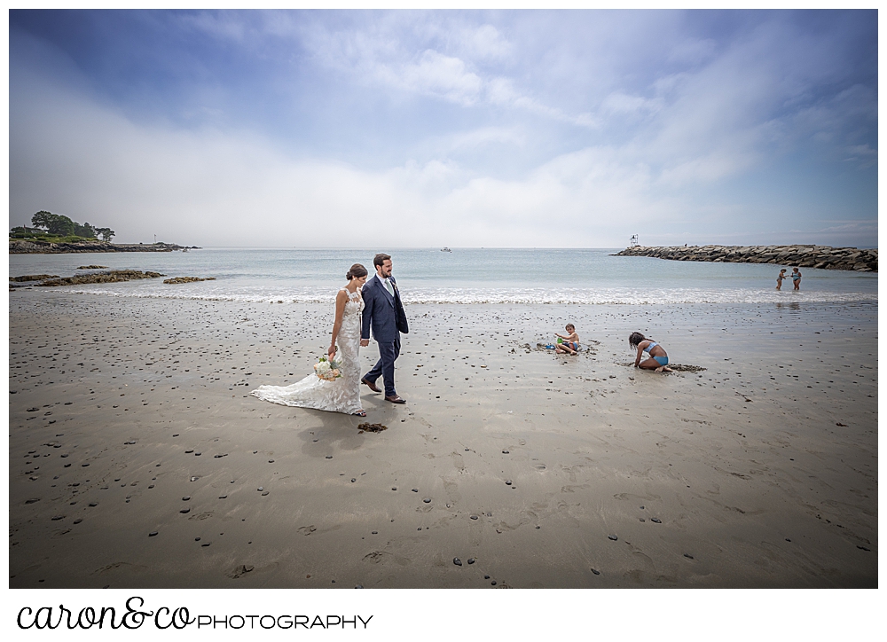 a bride and groom walk on Colony Beach in Kennebunkport, Maine, while children play in the sand