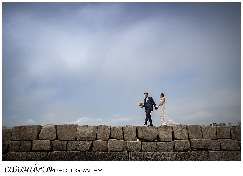 a bride and groom walk on the top of the Kennebunkport Breakwater, the groom is holding the bride's bouquet