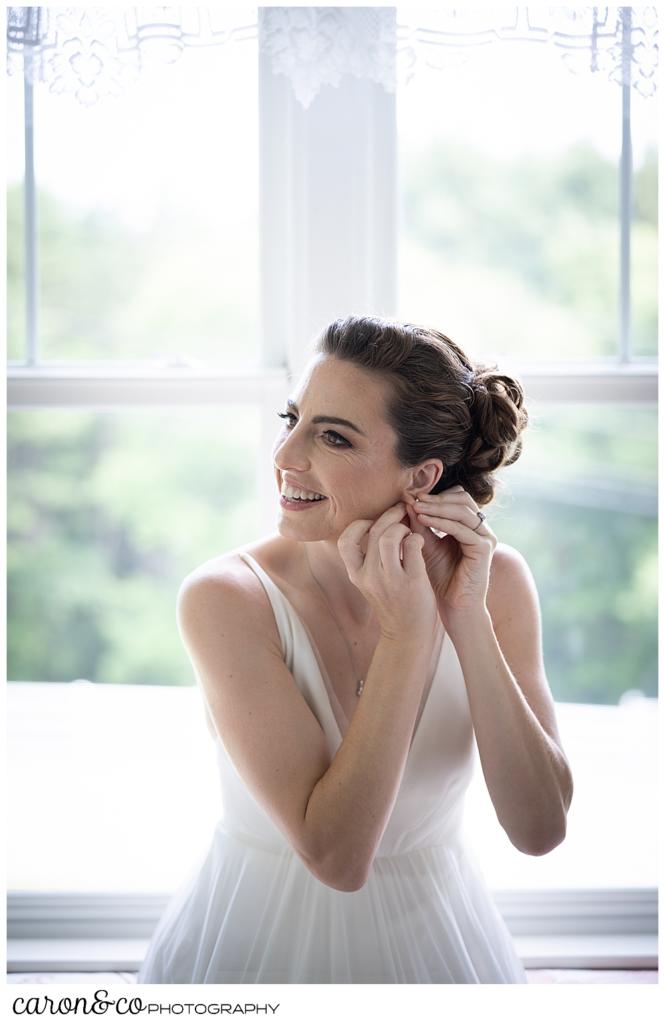 a bride, seated in a window seat, puts on an earring