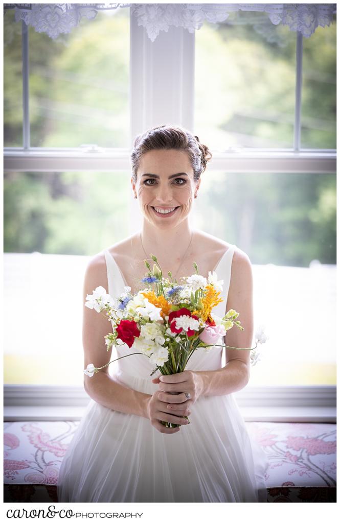 a smiling bride seated on a window seat, holding a bright colored bouquet of wildflowers