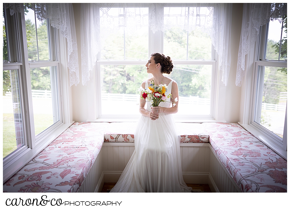 a bride sits on an alcove window seat, holding a bright colored wildflower bouquet, her head is turned as she looks out a window