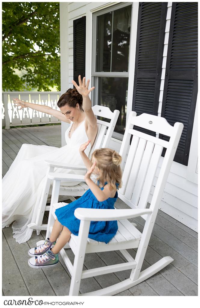 a bride, wearing a sleeveless white dress, sits on a porch in a white rocking chair, next to a small girl wearing a turquoise dress, they have their hands up in the air