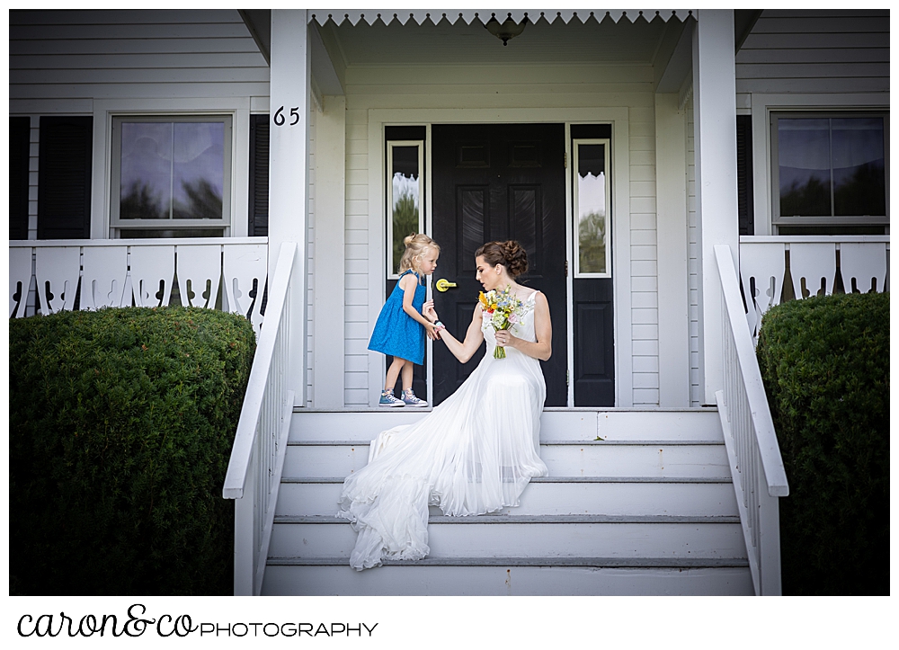 a bride in a sleeveless white dress, sitting on the front steps of a White House with black trim, as a small girl in a turquoise dress touches her wrist