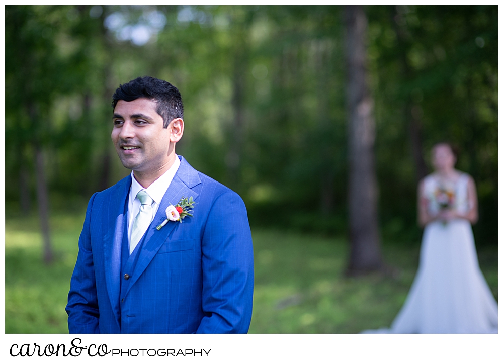 a groom in a blue suit stands with his back to his bride, seconds before their Maine wedding day first look