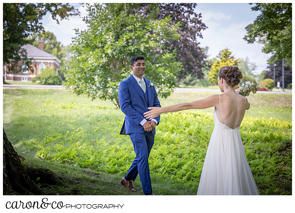 a bride in a sleeveless white dress holds her arms out to her groom wearing a blue suit, who is approaching her during their Maine wedding day first look