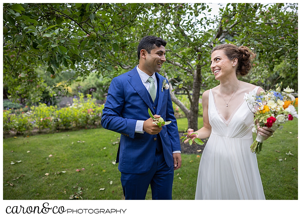 a bride and groom smile at each other while they each hold an apple, at their joyful Pineland Farms wedding