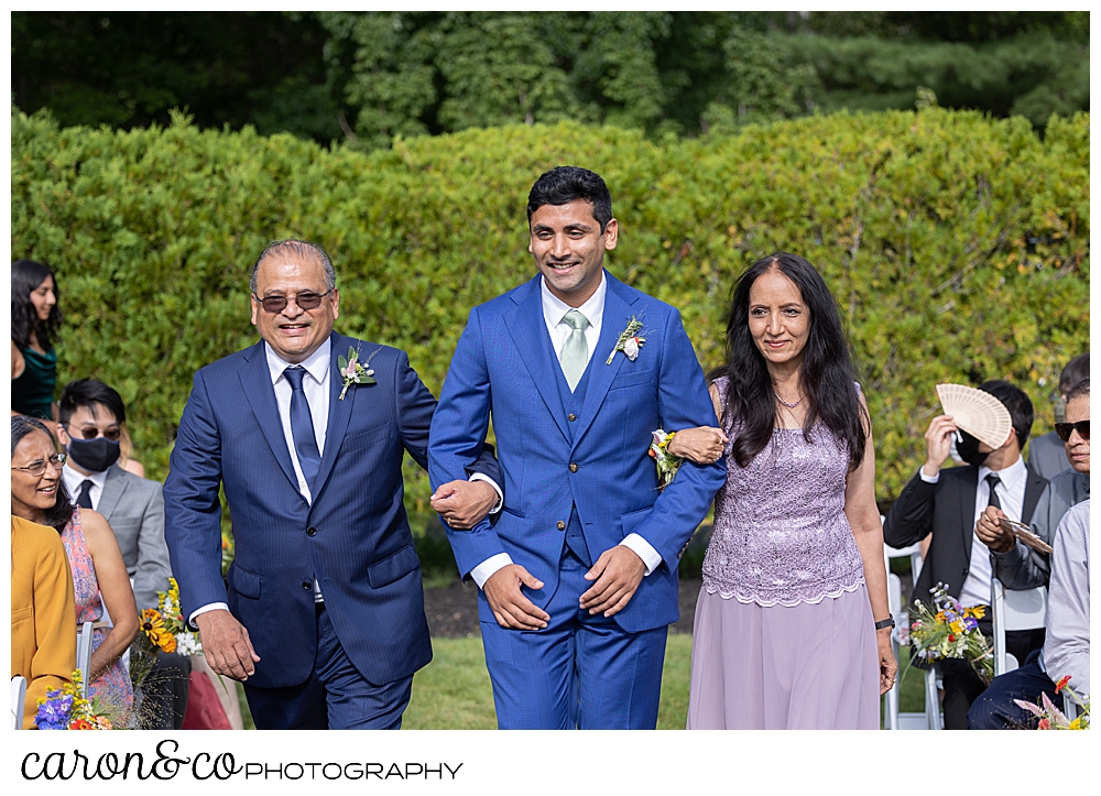 a groom walks down the aisle at the Pineland Farms Garden, with his mother and father