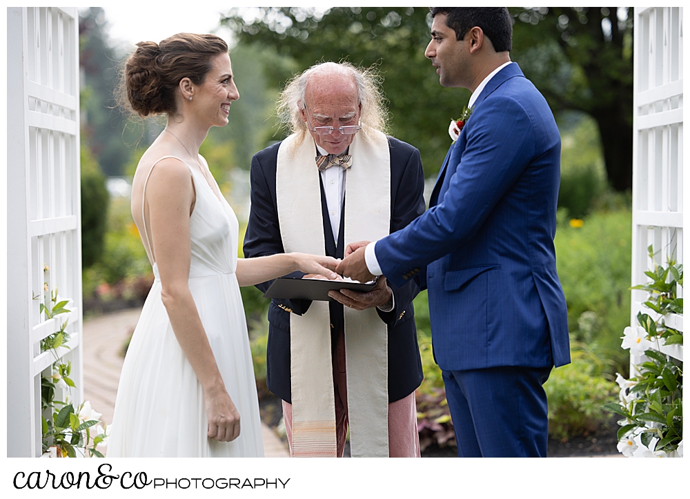 a groom wearing a blue suit, puts a wedding band on a bride wearing a sleeveless white dress, at a joyful Pineland Farms wedding ceremony