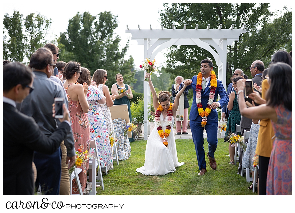 a bride walks down the Pineland farms garden aisle next to her groom, and she bends her knee low to the ground, with her arms up over her head