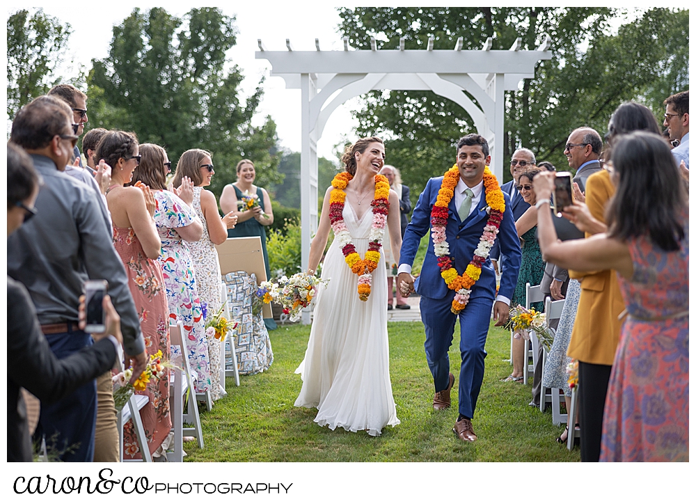 a bride and groom with colorful leis, smile during their recessional at a joyful Pineland farms wedding ceremony