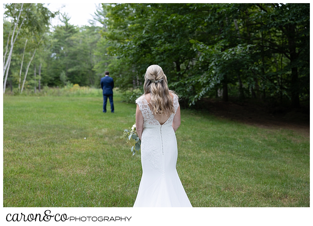 a bride is standing in the foreground with her back to the camera, while her groom is in the background, with his back turned as they prepare for their Kingsley pines maine wedding day first look