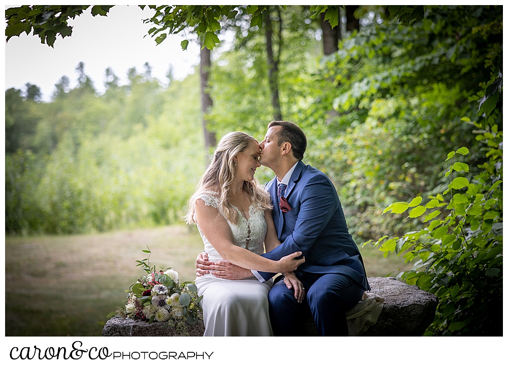 a bride and groom are sitting side by side on a stone bench, the groom is kissing the bride on her forehead