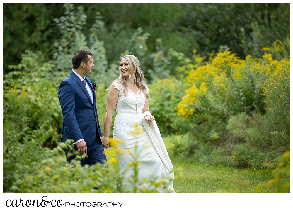 a bride and groom are walking side by side, holding hands, in a field at Kingsley Pines, Raymond, Maine