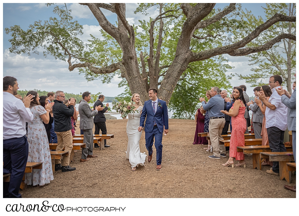 a bride and groom are holding hands and smiling during their Kingsley Pines Maine wedding ceremony recessional