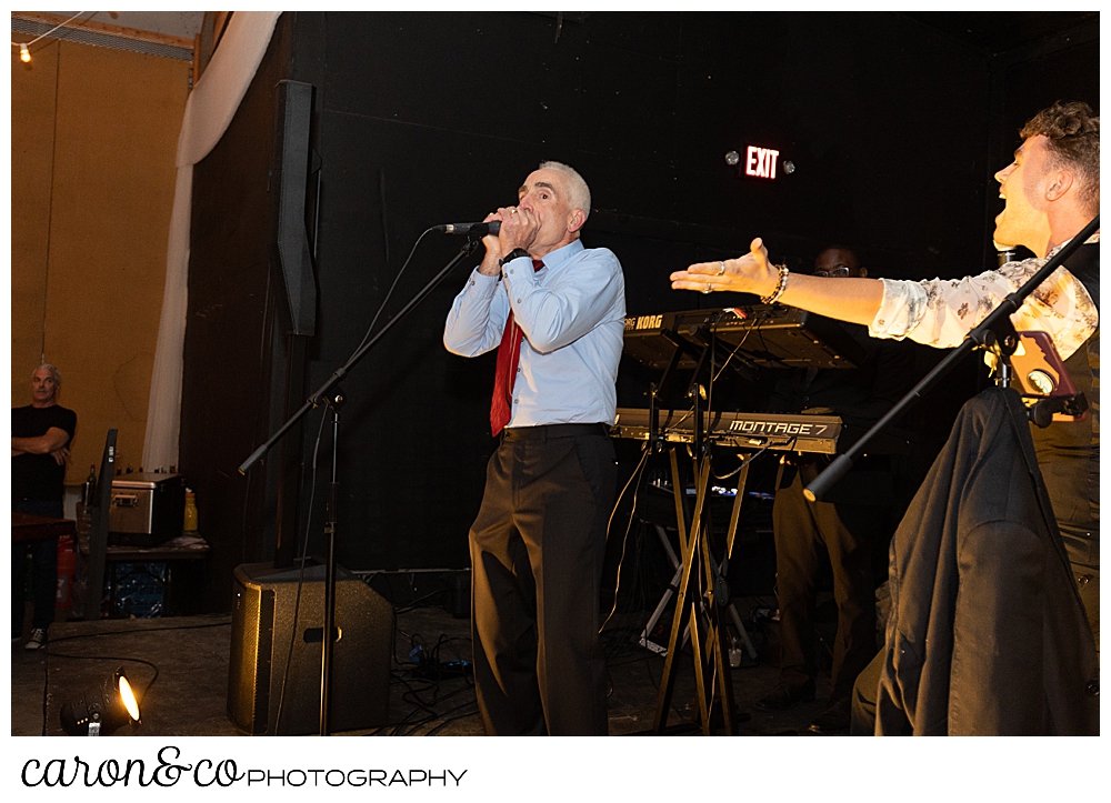 a man playing a harmonica on a stage, a band member holds a hand out towards the man playing the harmonica