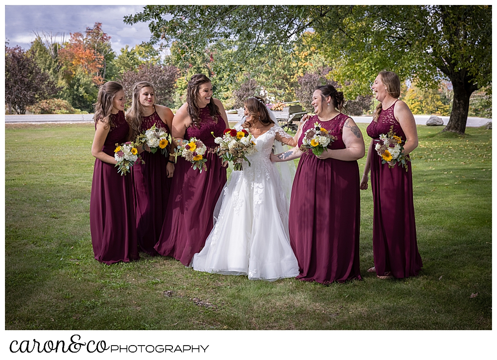 a bride wearing a white bridal gown, and her bridesmaids wearing Burgundy dresses, stand together in a line, laughing