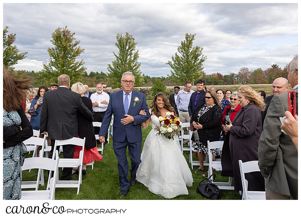 a bride wearing a white wedding dress, walks down the aisle on the arm of her dad wearing a blue suit at a wedding at Maine wedding Barn