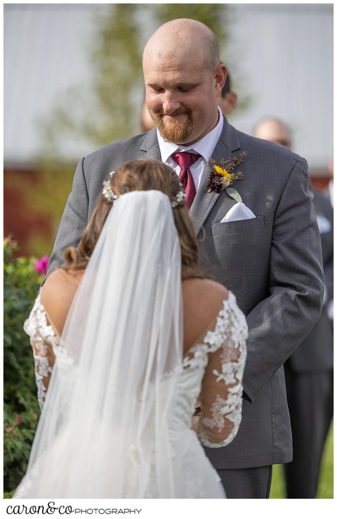 a bride wearing a white dress and veil with her back to the camera is in the foreground, her groom, wearing a gray suit is in the background smiling