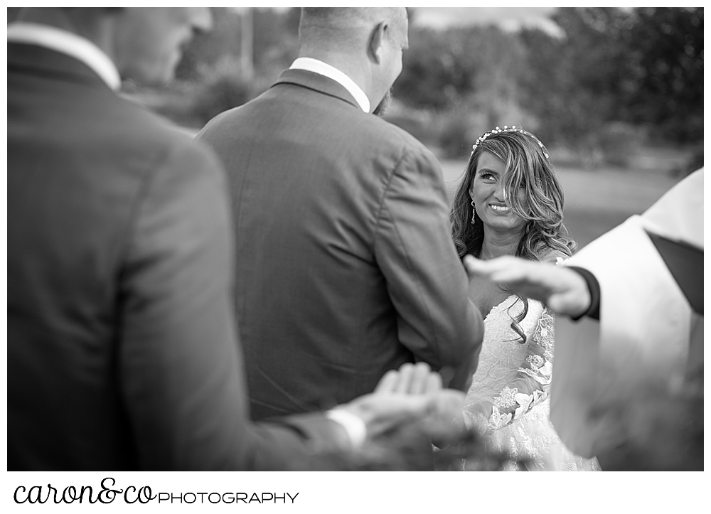a black and white photo of an officiant in the foreground, blessing the rings, and the bride smiling at her groom in the background