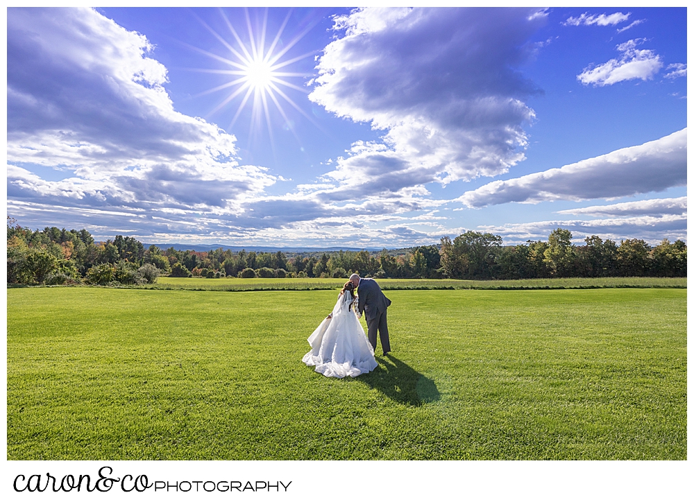 a bride and groom kiss in a grassy field, with clouds and a sun burst overhead