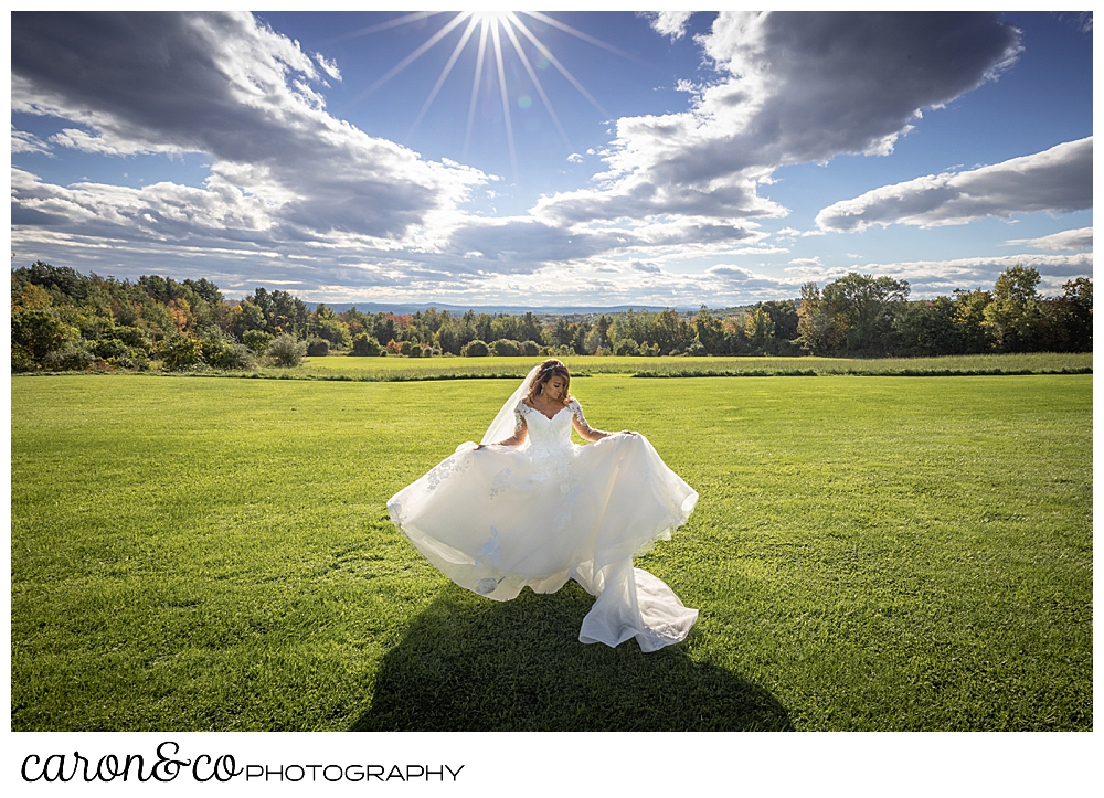 a bride holds the skirt of her wedding dress, as she twirls in a grassy field at a wedding at Maine wedding barn in Minot Maine