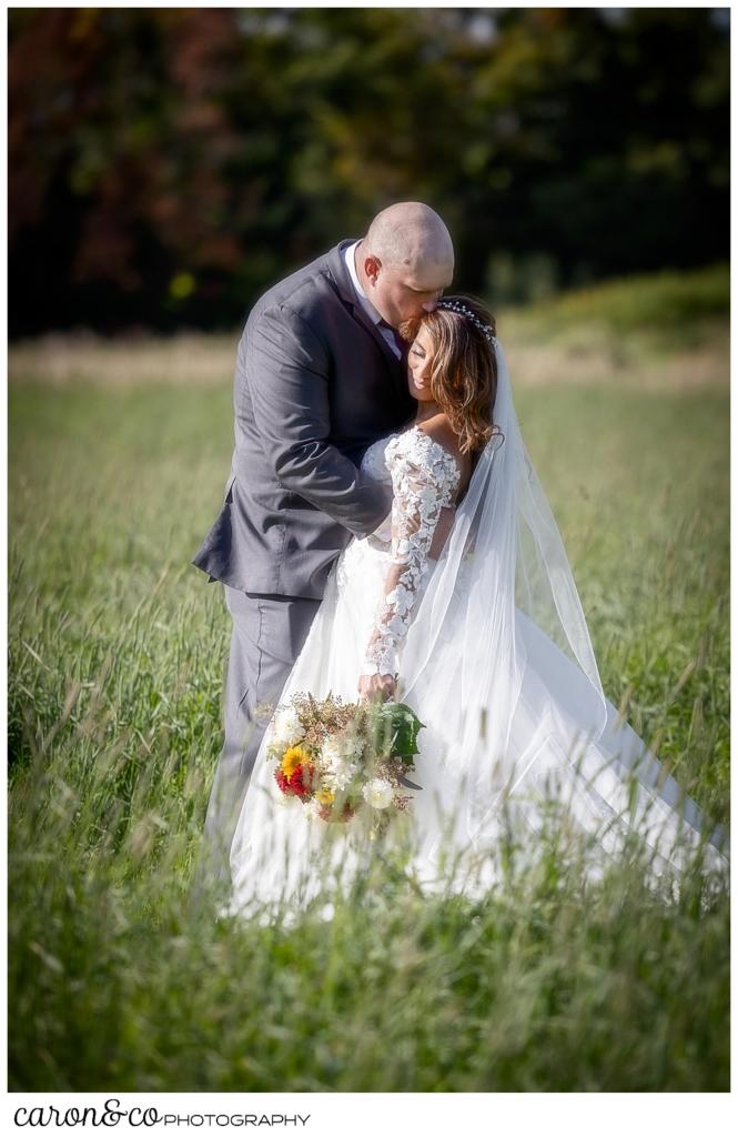 a groom kisses a bride's cheek as they stand face to face, in a field of grass