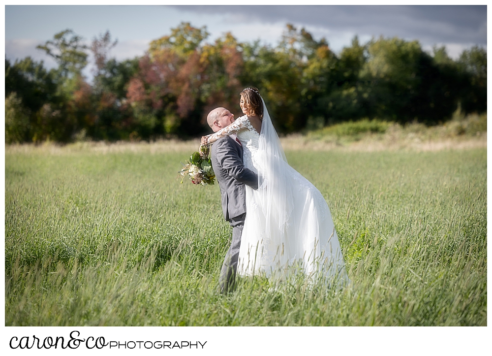 a groom lifts his bride in a grassy field at the Maine wedding barn in Minot Maine