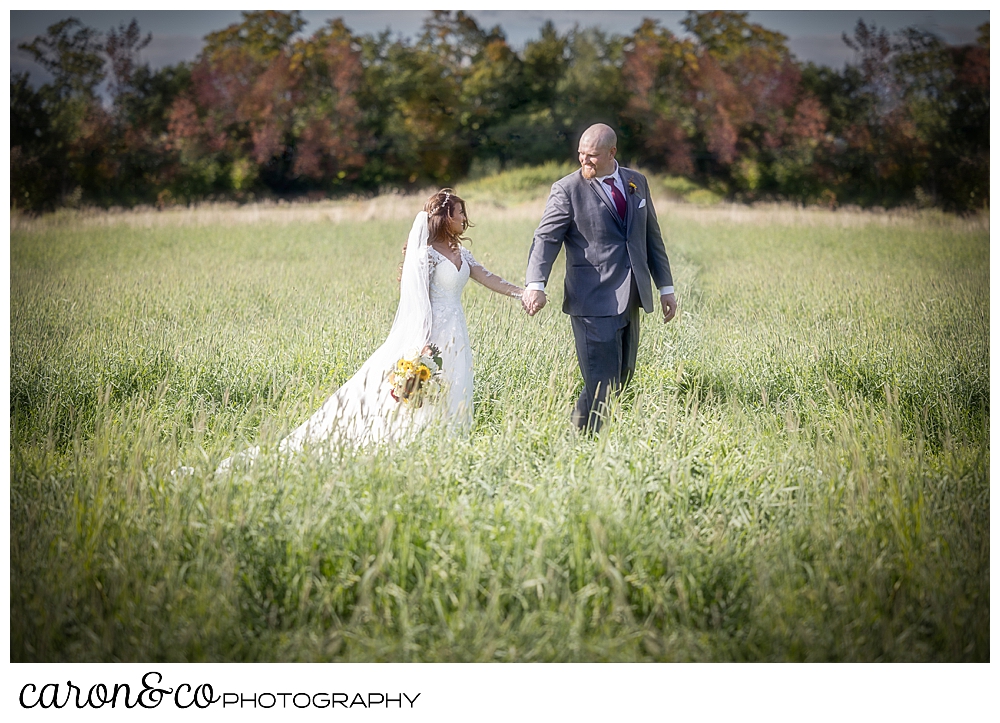 a bride and groom hold hands and walk through a field, the groom is leading the bride