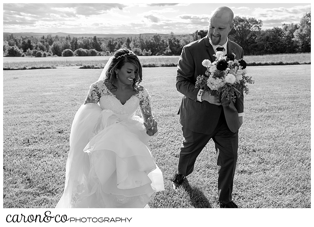 black and white photo of a bride and groom walking together, the groom is carrying the bride's bouquet, and the bride is looking at her wedding band